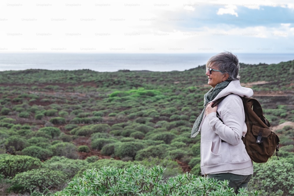 Mujer mayor sonriente disfrutando de una excursión al aire libre entre arbustos verdes y el mar. Ancianos de pelo gris con mochila mirando al horizonte