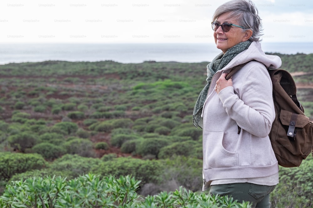 Lächelnde ältere Frau, die einen Ausflug im Freien zwischen grünen Büschen und Meer genießt. Grauhaarige ältere Menschen mit Rucksackliebhaber der Natur