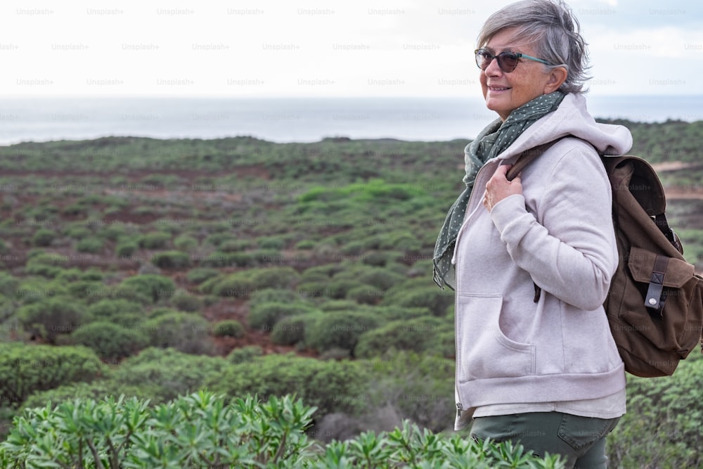 Donna anziana sorridente che si gode un'escursione all'aperto tra cespugli verdi e mare. Anziani dai capelli grigi con zaino amante della natura