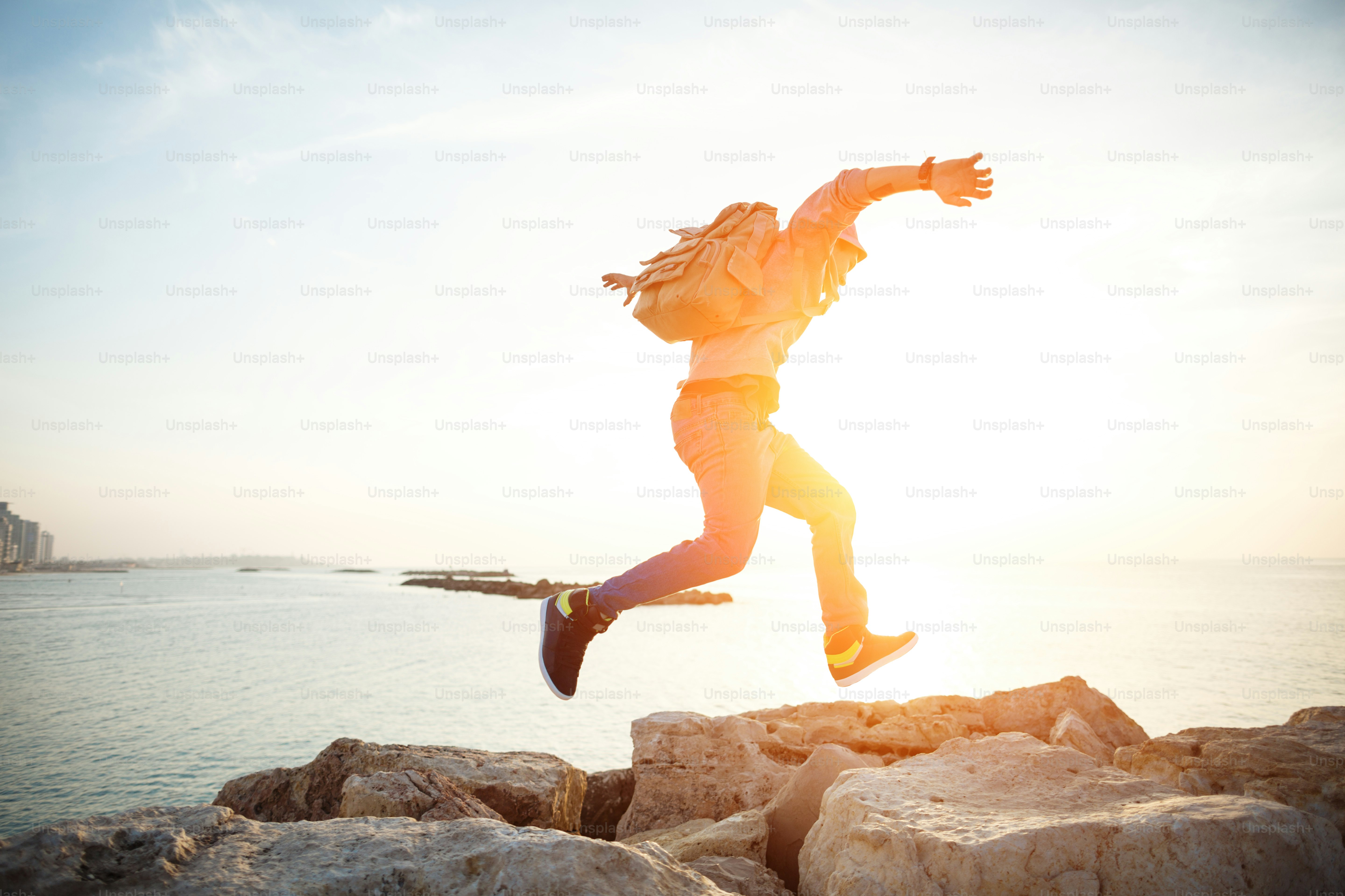 Brave man with backpack running over rocks near ocean in sunset photo ...