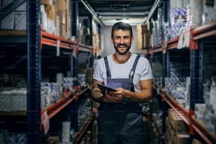 Smiling bearded tattooed hardworking employee holding tablet and standing in storage.