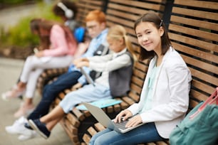 Asian schoolgirl typing on laptop computer and smiling at camera while sitting on the bench outdoors together with her classmates in the background