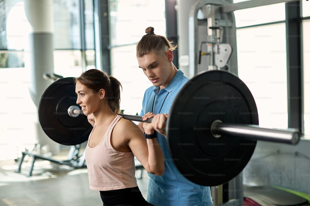 Junge Sportlerin hebt Langhantel, während sie mit ihrem Trainer in einem Fitnessstudio Krafttraining macht.