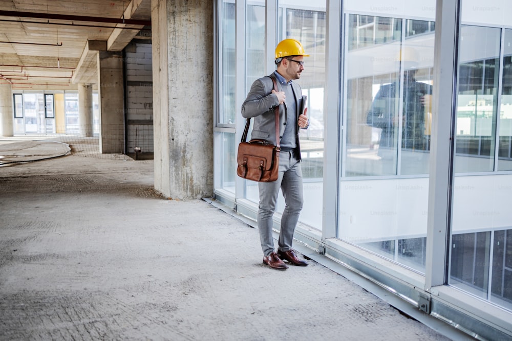 Young unshaven architect in suit with helmet on head standing in building in construction process and looking trough window.