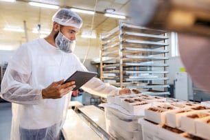 A food inspector in a sterile white uniform is holding the tablet and looking at collected cookies. Food check is important if we want quality and healthy food.