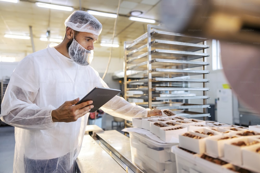 A food inspector in a sterile white uniform is holding the tablet and looking at collected cookies. Food check is important if we want quality and healthy food.
