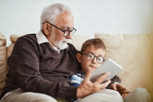 Grandfather and grandson using digital tablet while relaxing on the sofa at home.