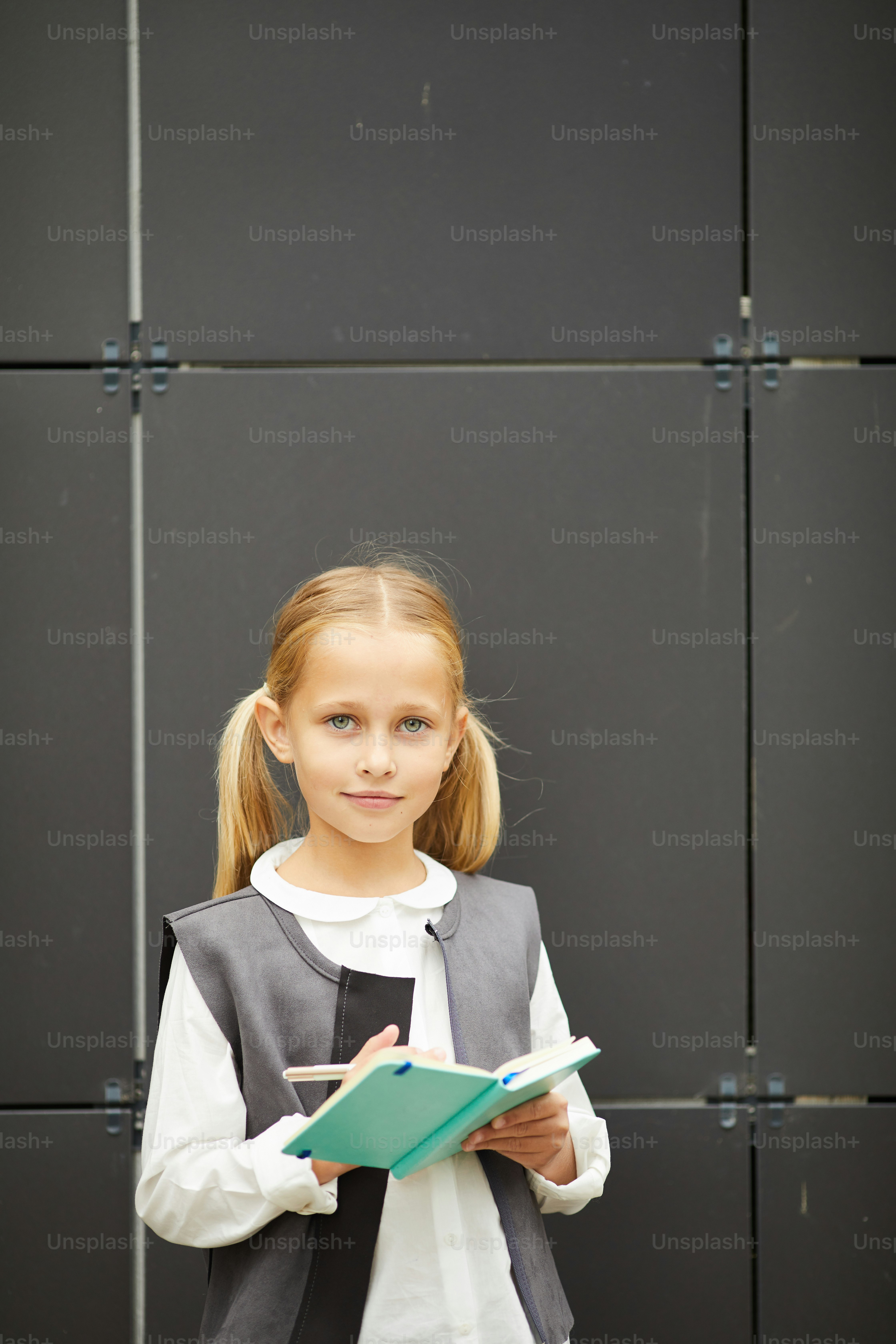 Portrait of cute schoolgirl with blond hair standing and looking at camera while making notes in her notepad outdoors