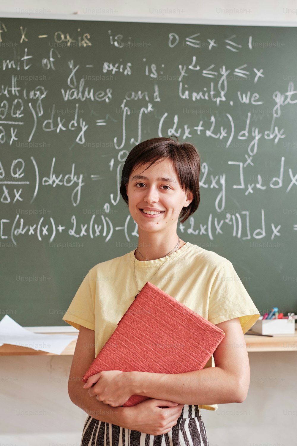 Portrait of teacher with books smiling at camera standing against the blackboard and teaching at university