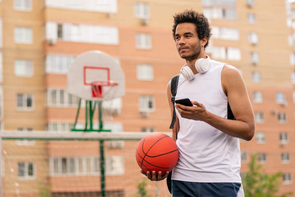 Young mixed-race athlete with ball and smartphone texting at break on playground in urban environment