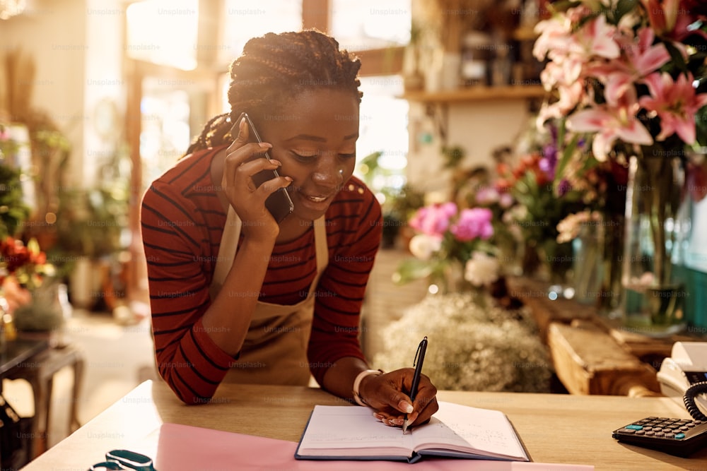 젊은 아프리카계 미국인 꽃집은 꽃집에서 주문을 받는 동안 휴대전화로 이야기하고 메모를 하고 있습니다.