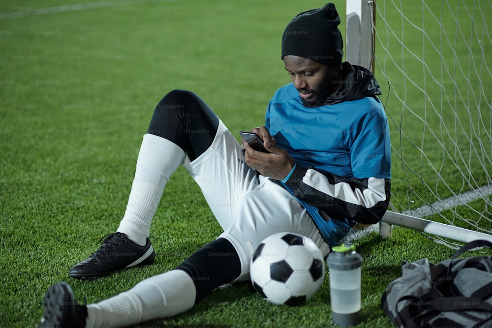 Young restful sportsman in uniform scrolling in smartphone after training while sitting on green lawn by net