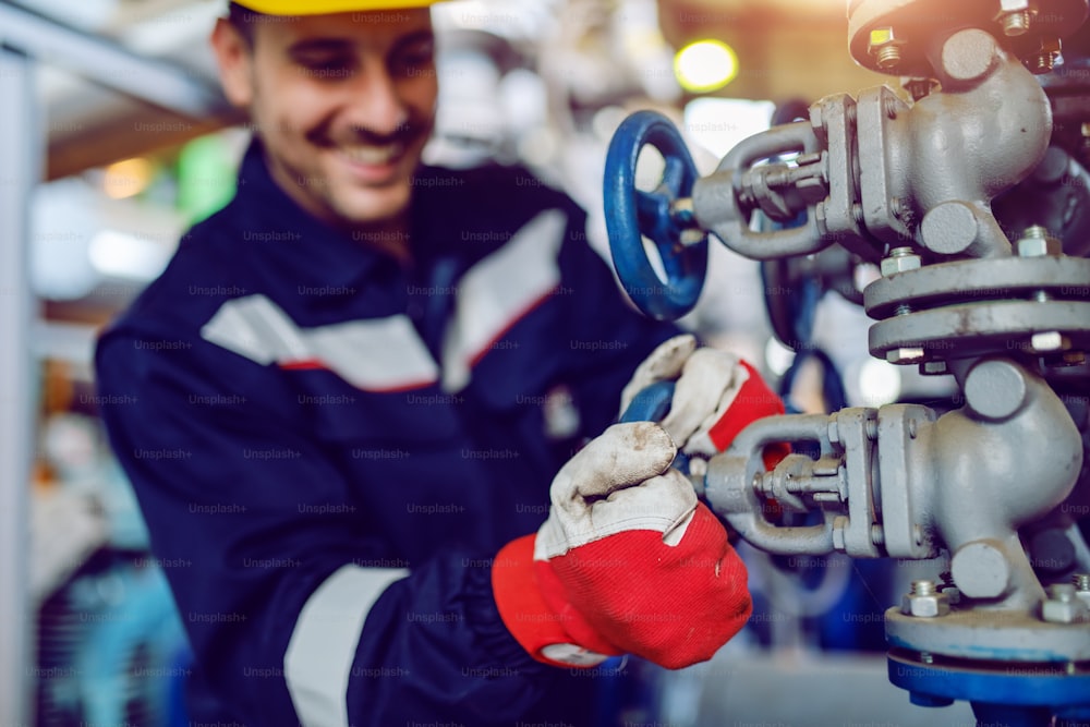 Smiling hardworking energy plant worker in working suit screwing valve. Selective focus on hands.