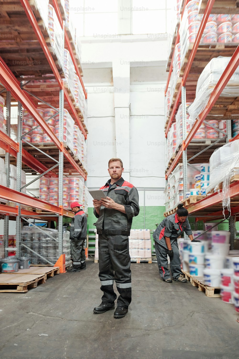 Serious young male manager of warehouse in uniform using digital tablet while standing in aisle between racks on background of colleagues