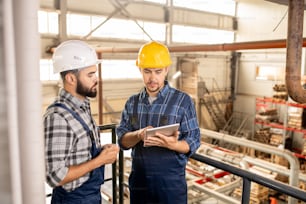 One of young contemporary engineers in helmets and workwear showing his colleague online information on tablet display