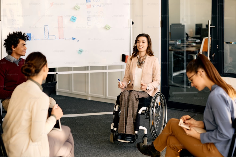 Feliz gerente ejecutiva en silla de ruedas explicando la estrategia comercial de sus compañeros de trabajo en la pizarra durante la presentación en la sala de reuniones.