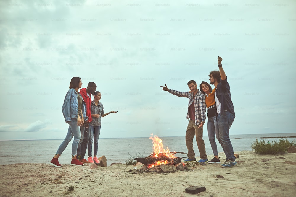 Get together. Happy friends standing near bonfire while telling stories each other at the sea shore