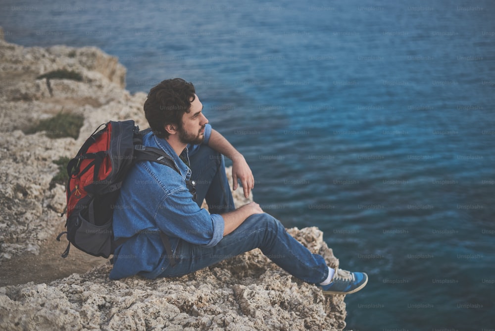 Il turista maschio calmo è seduto sulla roccia e si gode la vista sul mare. Porta lo zaino sulla schiena