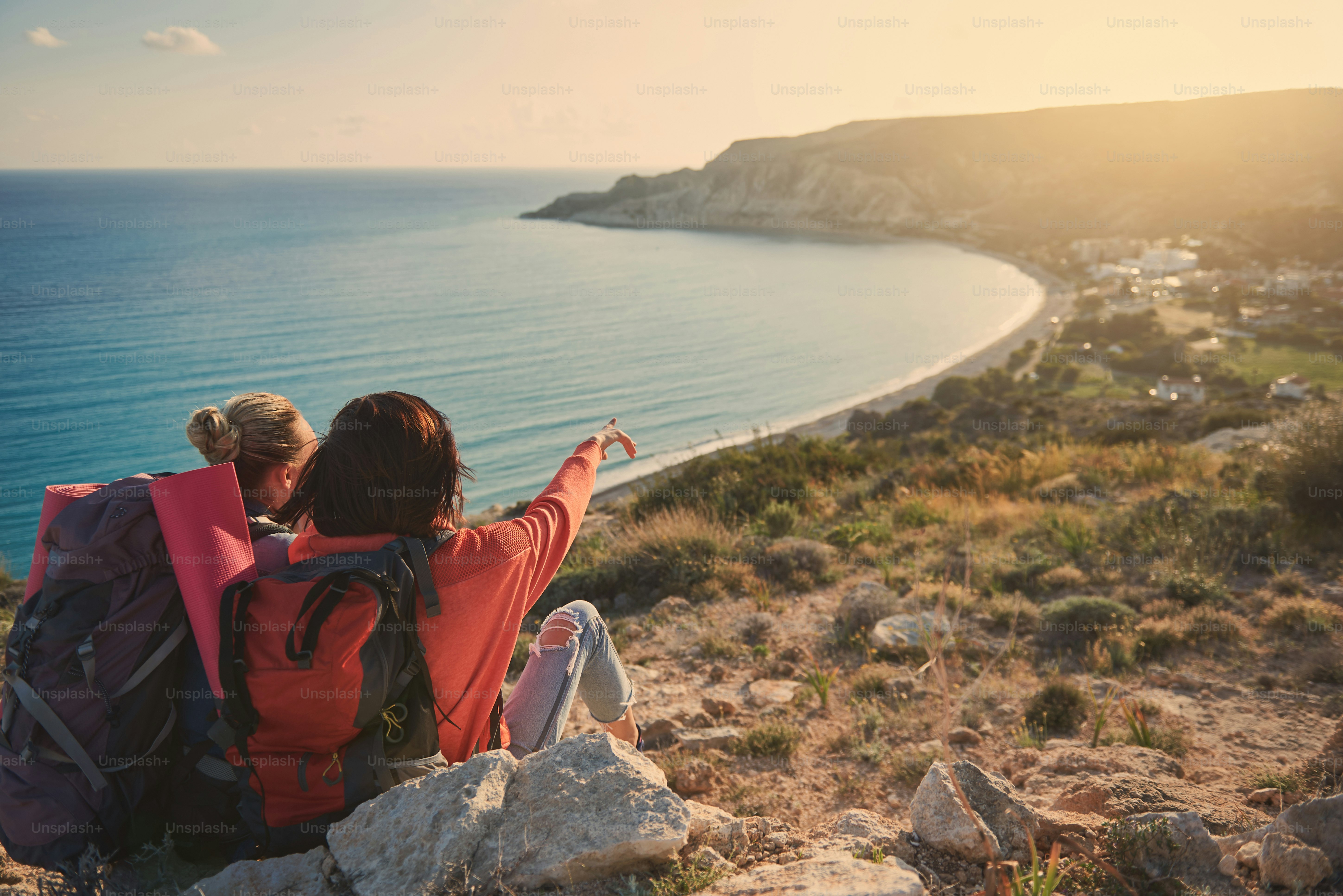 Girls with rucksacks sitting high with their backs and enjoying wonderful sea view. One of them is pointing. Copy space in right side