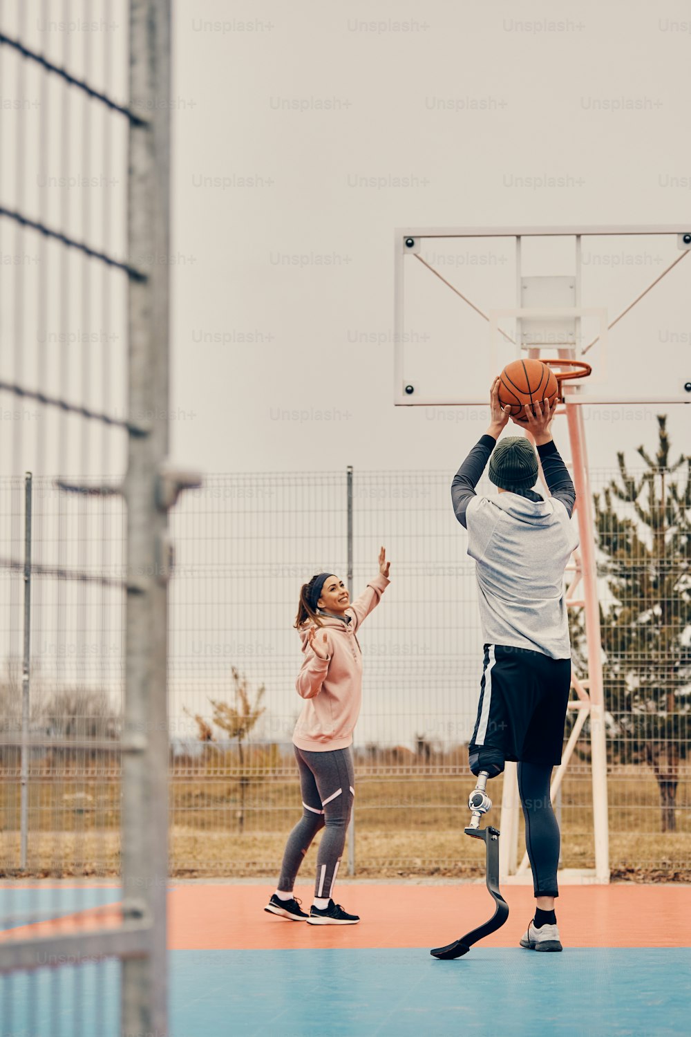 Back view of sportsman with leg prosthesis playing basketball with female friend and shooting at the hoop on outdoor sports court.