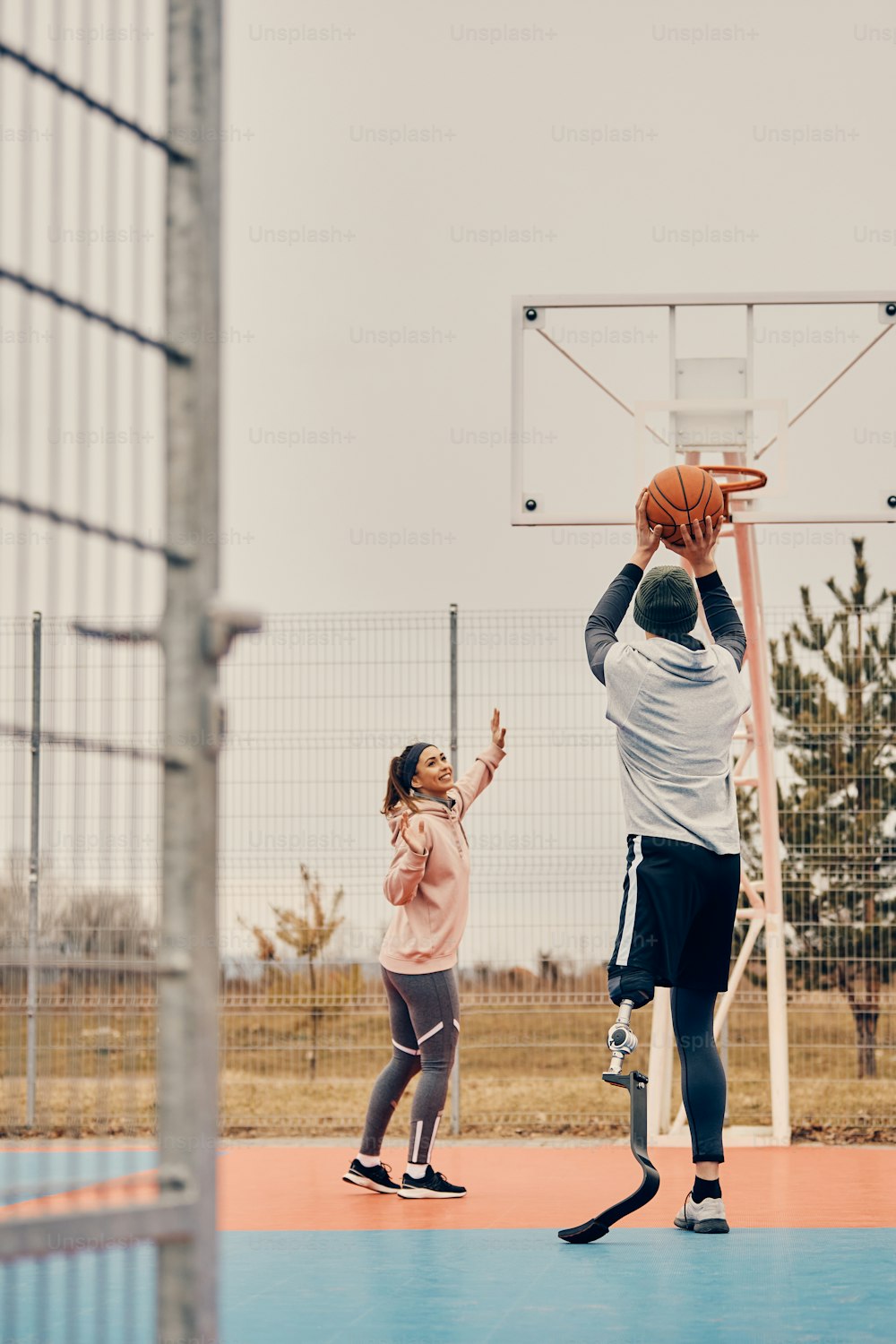 Vista posterior de un deportista con prótesis de pierna jugando al baloncesto con una amiga y disparando al aro en una cancha deportiva al aire libre.