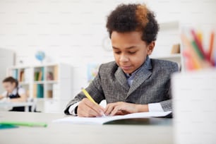Niño africano sentado en el escritorio y tomando notas en su cuaderno durante la lección en la escuela