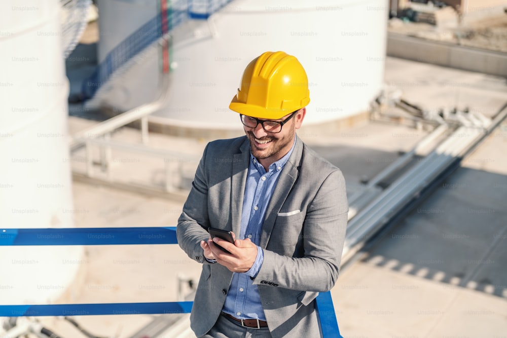 Young smiling successful attractive elegant rich businessman in suit with helmet on head leaning on the railing and using smart phone. Refinery exterior.