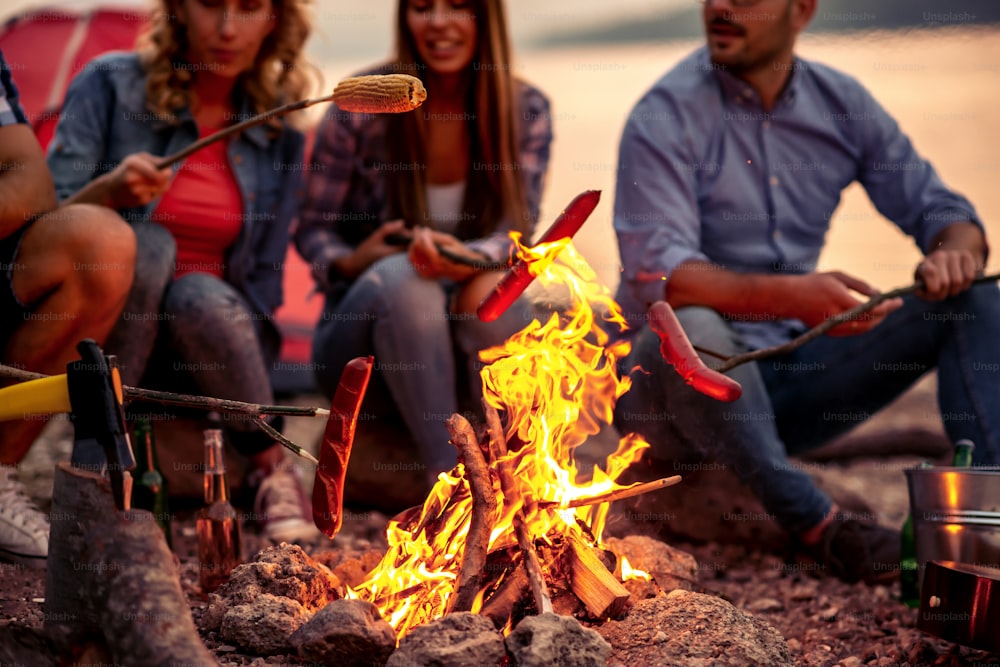 Group of friends sitting around the fire on sunset, grilling sausages and having great time on the beach.