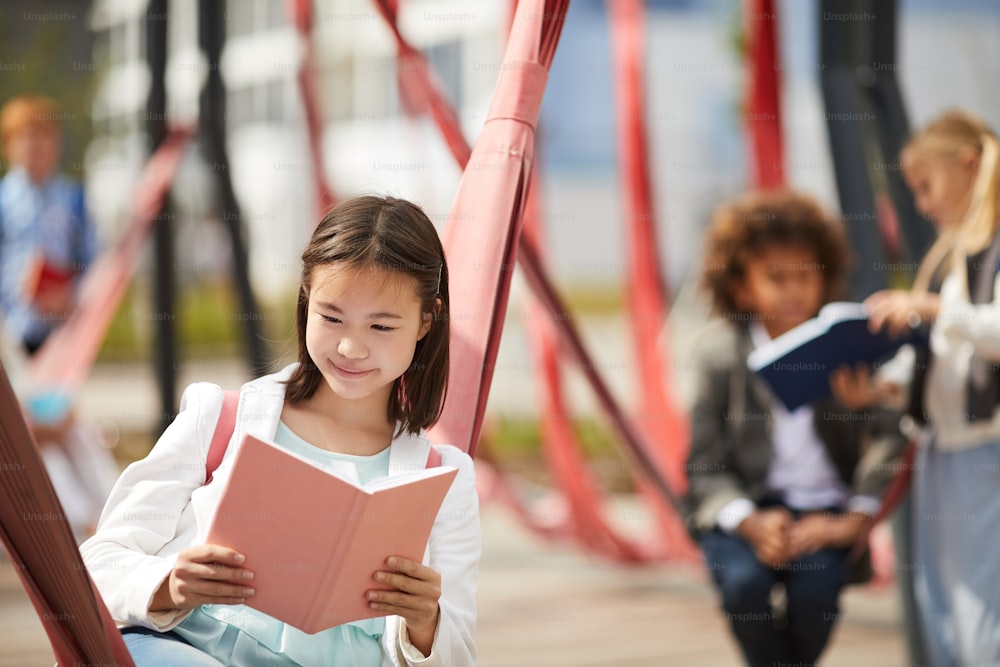 Asian schoolgirl reading a book outdoors on the school playground with her classmates in the background