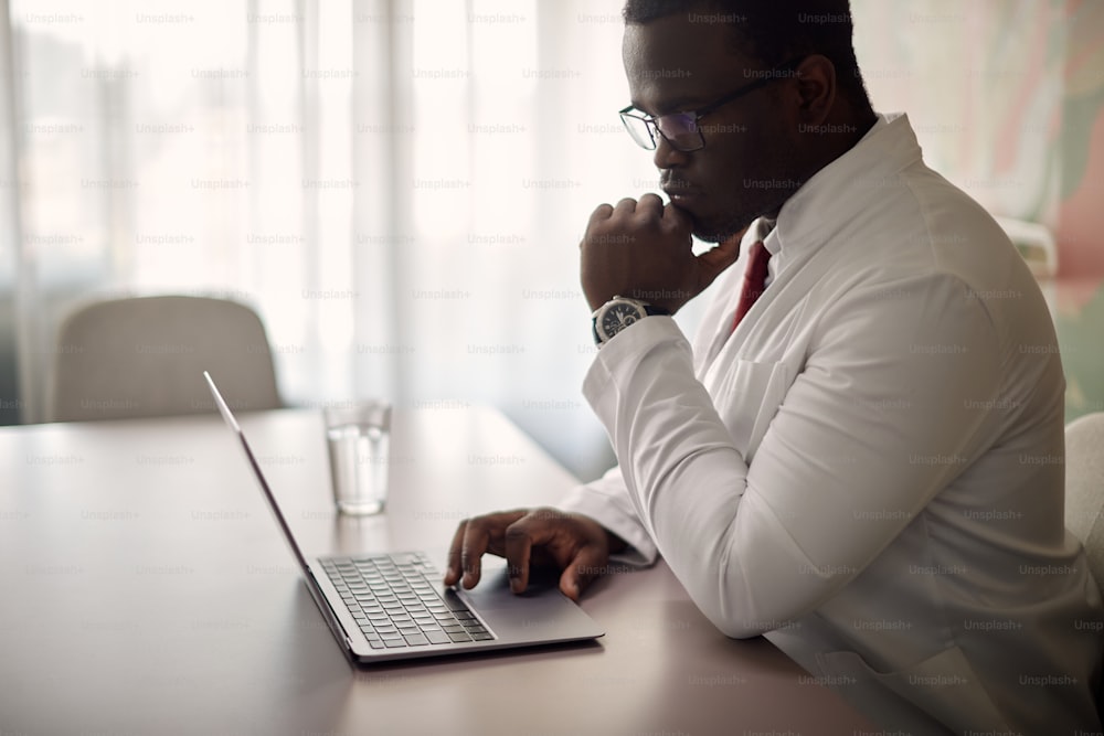 Nachdenklicher schwarzer Arzt mit Laptop während der Arbeit in seinem Büro in der Klinik.