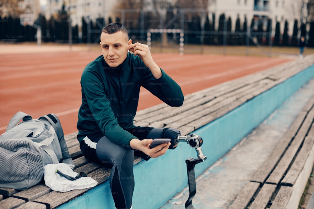 Joven atleta con discapacidad en las piernas escuchando música con auriculares mientras se relaja en un banco en el campo de deportes al aire libre.