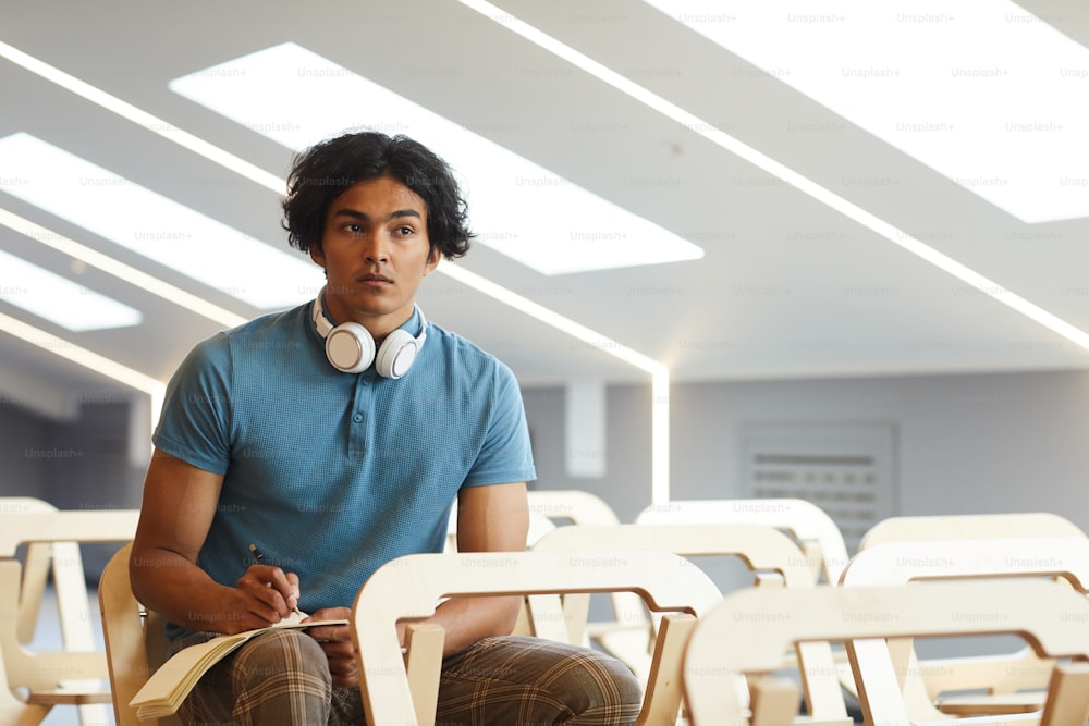 Concentrated mixed race guy with headphones around neck sitting in empty auditorium and making notes in workbook