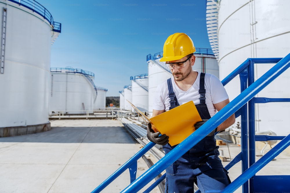 Hardworking handsome caucasian worker in overall and with helmet on head standing on the stairs and looking at folder with documents. Oil production.