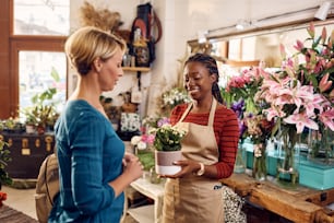 Happy African American florist assisting her customer in buying potted plant at flower shop.