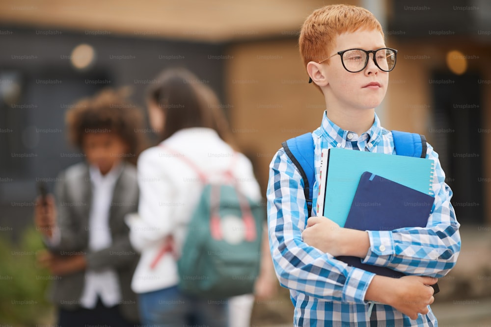 Schoolboy on eyeglasses holding books and notebooks while standing outdoors with his classmates in the background