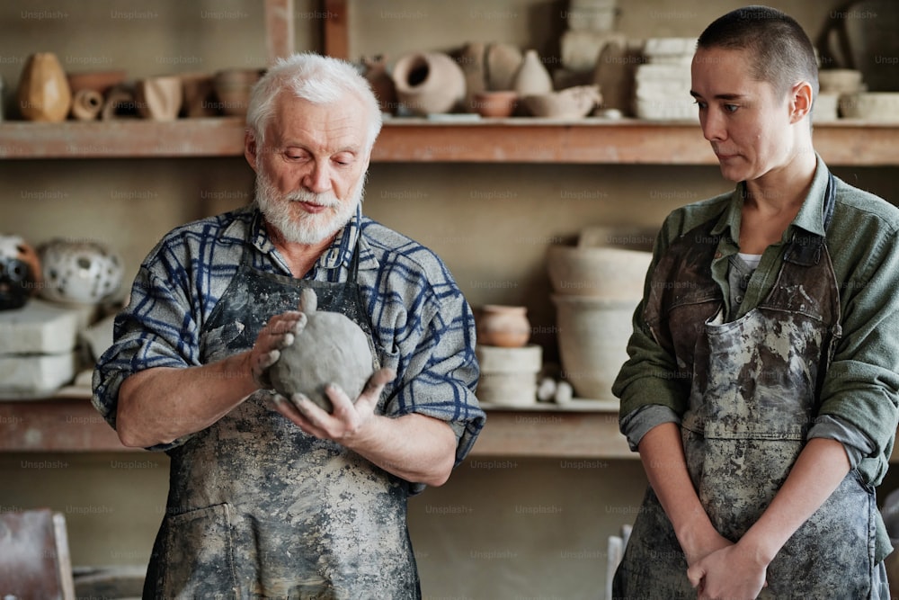 Senior man in apron turning a piece of clay in his hands and teaching to his assistant of pottery craft