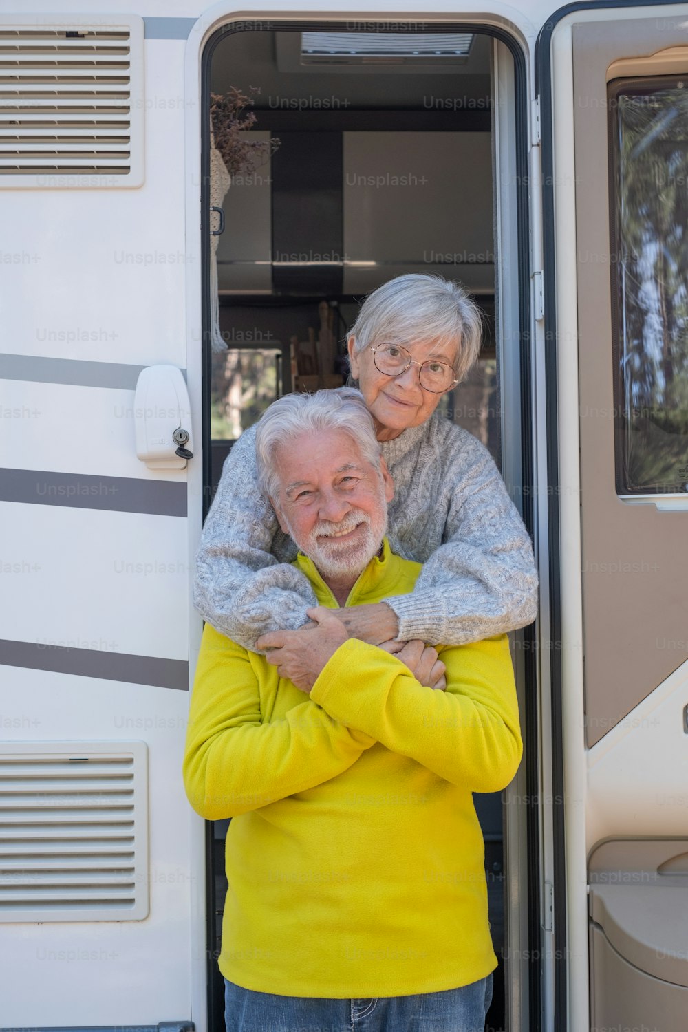 Happy relaxed caucasian senior couple on the door of their camper van motor home embrace looking at camera. Smiling attractive elderly people enjoying freedom vacation travel in the forest.