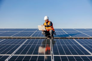 A handyman looking at the laptop on rooftop and testing solar panels.