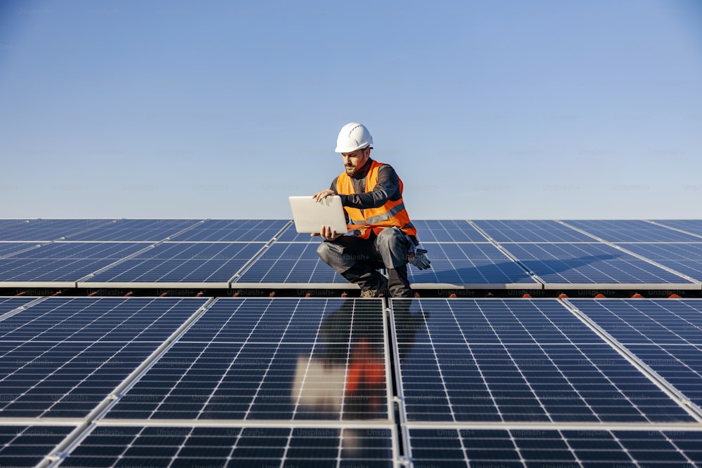 A handyman looking at the laptop on rooftop and testing solar panels.