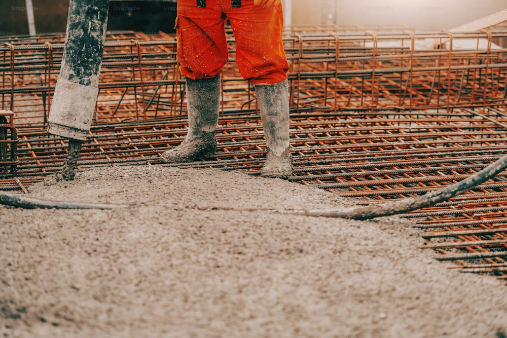 Construction site worker in boots and orange protective suit pouring concrete.