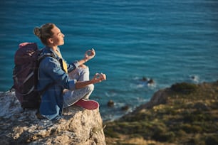 Profile of quiet girl taking her seat in lotus pose on high. Her eyes are closed. Coastline on background. Copy space in right side