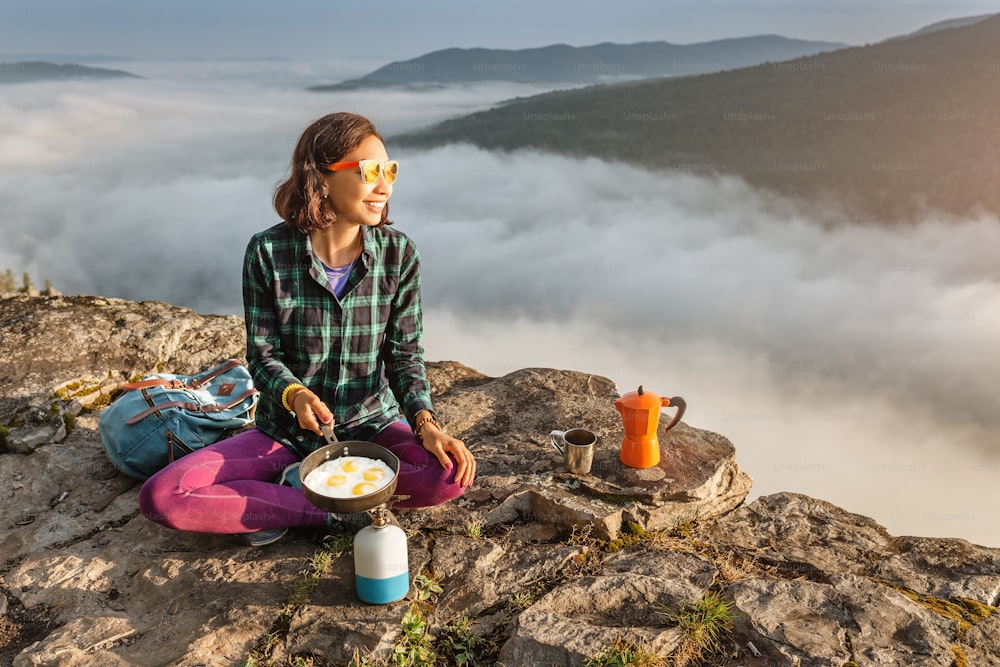 A woman traveler cooking and eating breakfast scrambled eggs and coffee at dawn in the misty mountains. Camping equipment and adventure concept