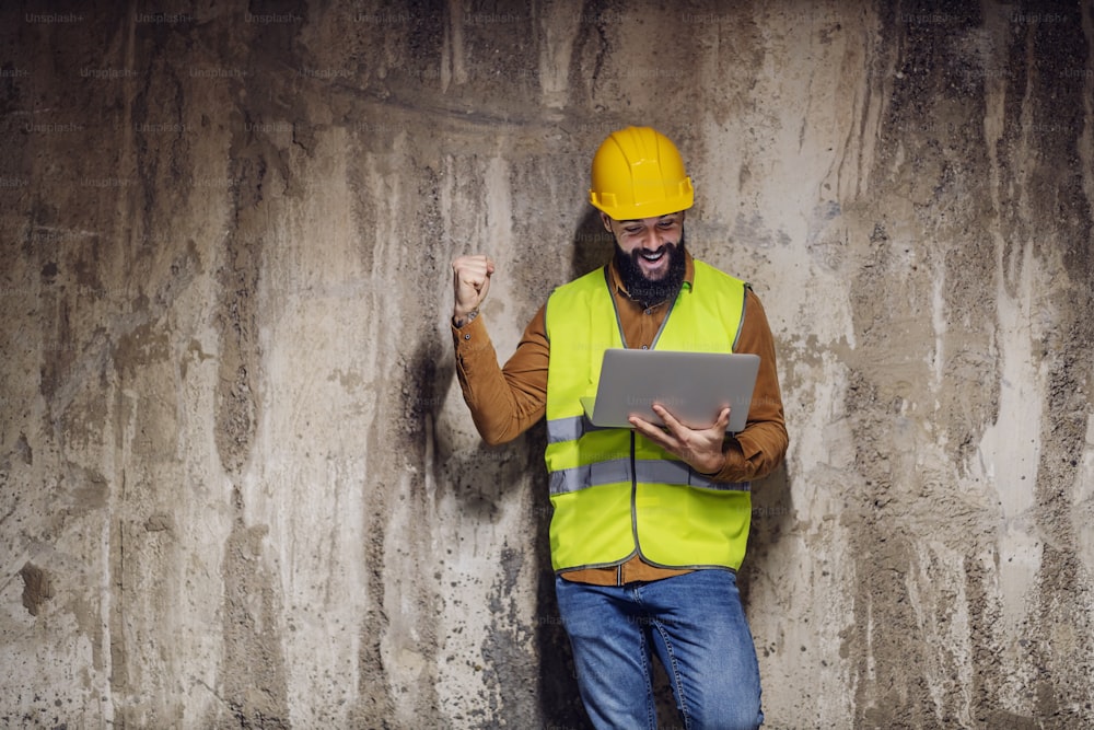 Three quarter length of cheerful handsome bearded supervisor in vest with safety helmet on head leaning on concrete wall. holding laptop and celebrating success.