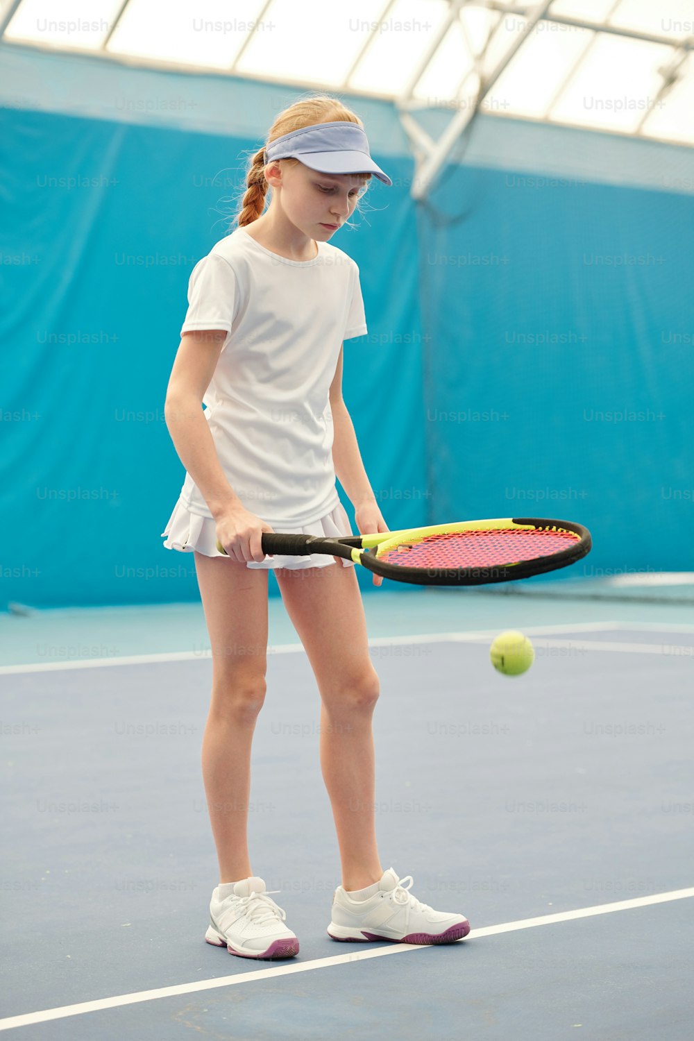 Youthful blond girl in white activewear holding tennis racket while standing on stadium and pushing ball during training in front of camera