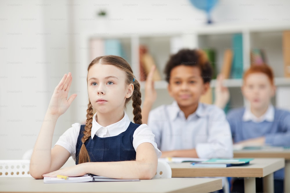 Serious schoolgirl sitting at desk and raising her hand with other classmates in the background during a lesson at school