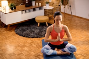 Vista de ángulo alto de mujer atlética meditando con los ojos cerrados mientras está sentada en postura de namaste durante el entrenamiento en casa.