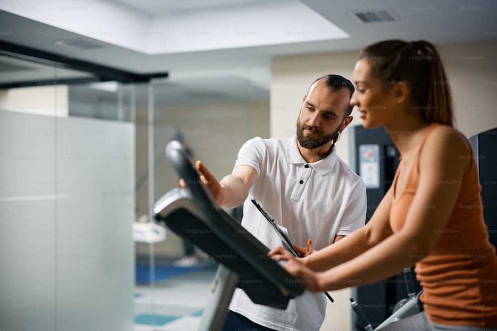 Athletic woman cycling on exercise bike while personal trainer is assisting her during sports training at health club. Focus is on a coach.