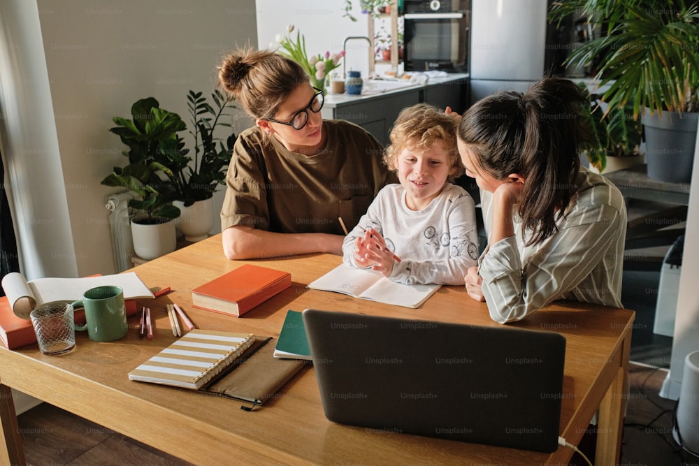 Two homosexual mothers helping son with study, they sitting at table with laptop and teach him online