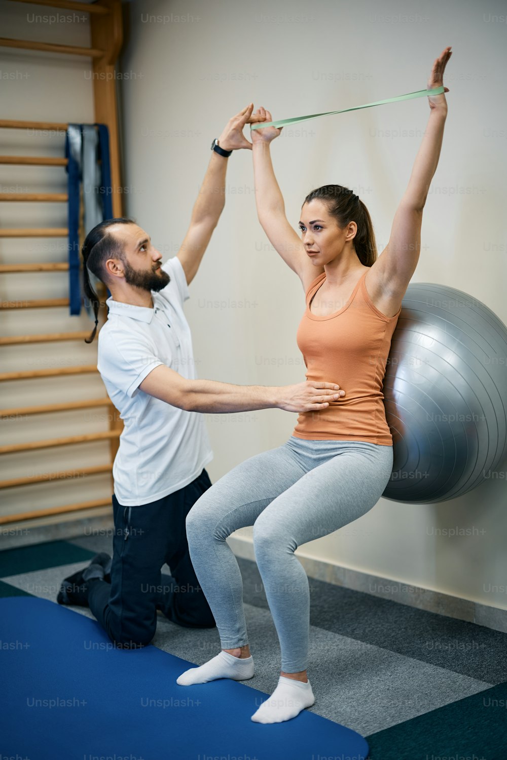 Fisioterapeuta que auxilia jovem esportista que está se exercitando com bola de fitness e banda de resistência durante o tratamento de fisioterapia no health club.