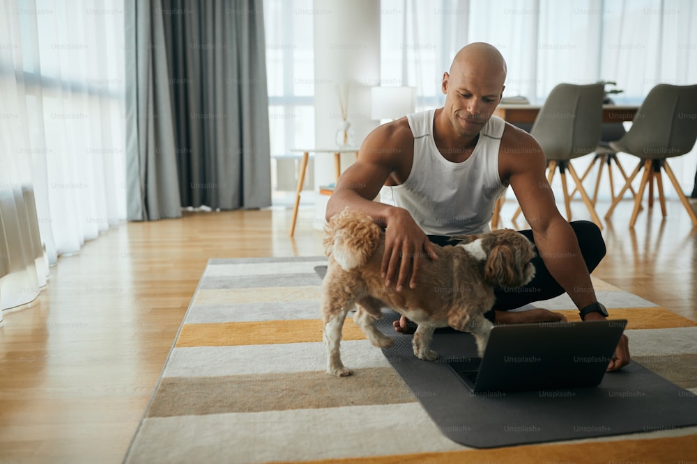 Happy African American athlete cuddling his dog while using laptop in the living room. Copy space.